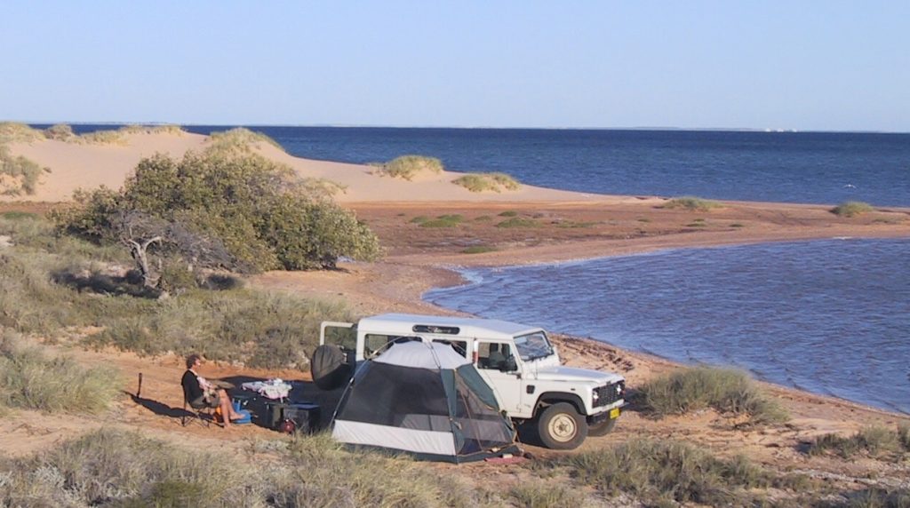 A white Land Rover parked next to a tent on a sandy beach, with a person sitting in a chair overlooking a deep blue ocean under a clear sky.